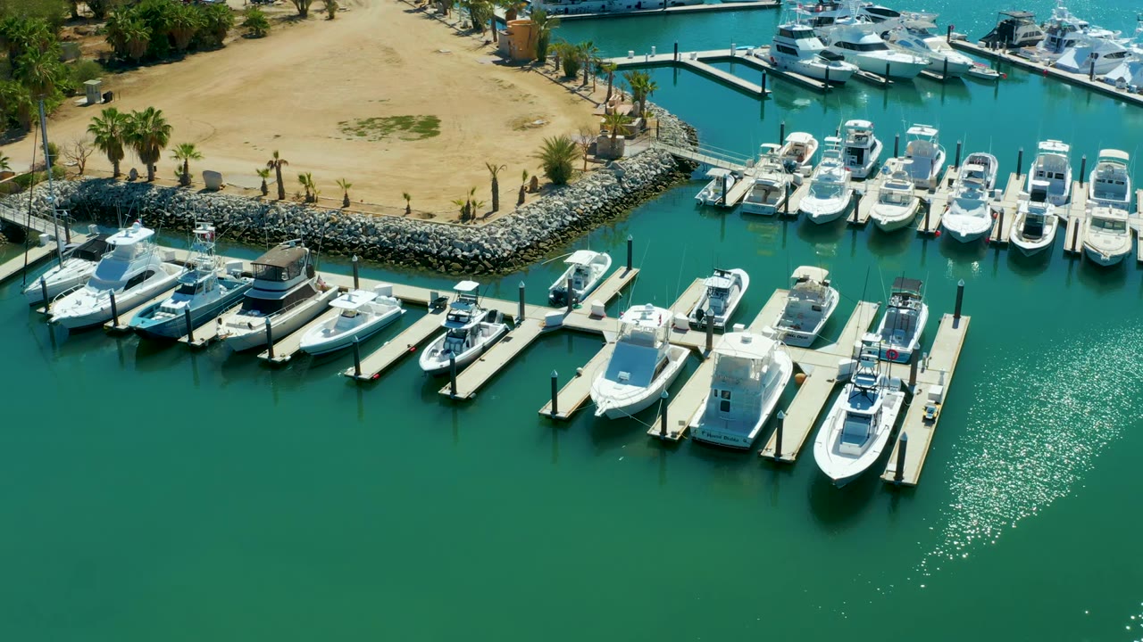 Puerto Los Cabos marina from above with rows of docked white boats, bright reflection on the turquoise water, sandy land and palms at the top of frame, and no visible people.