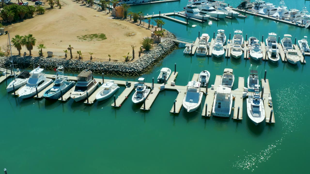 Puerto Los Cabos marina from above, with white boats packed along slips, a sandy bank and palms at the top, open rippled water below, and no visible people.