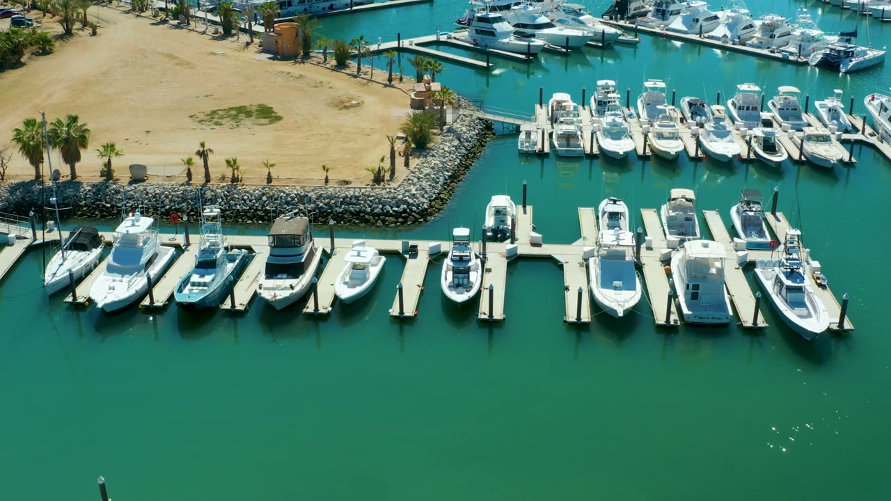 Opening aerial frame over Puerto Los Cabos marina with white boats tied to docks, green-blue water below, sandy shore and palms at the top, and no visible people.