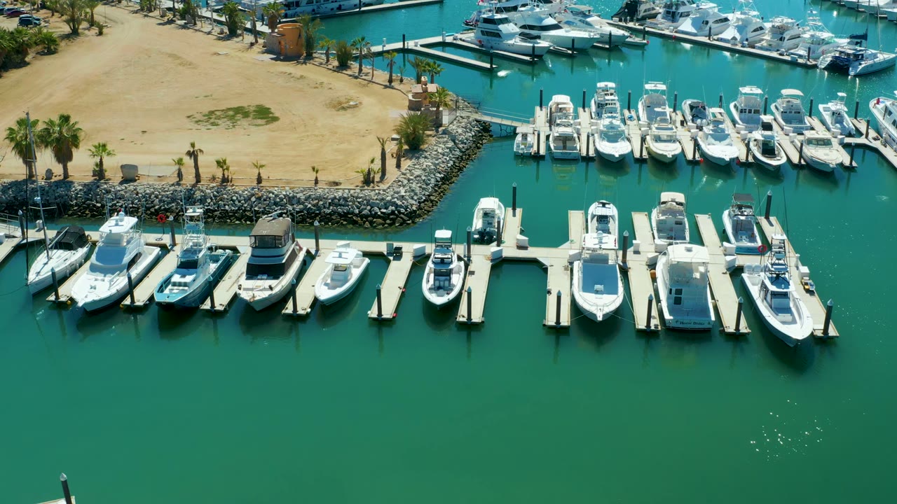 Mid-clip aerial frame at Puerto Los Cabos showing a row of white boats in marina slips, broad green water in the foreground, sandy land and palms at the top, and no visible people.