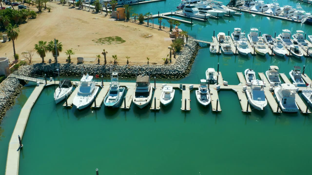 Opening aerial frame of Puerto Los Cabos marina, with white boats tied to docks, green-blue water below, sandy shore and palms above, and no visible people.