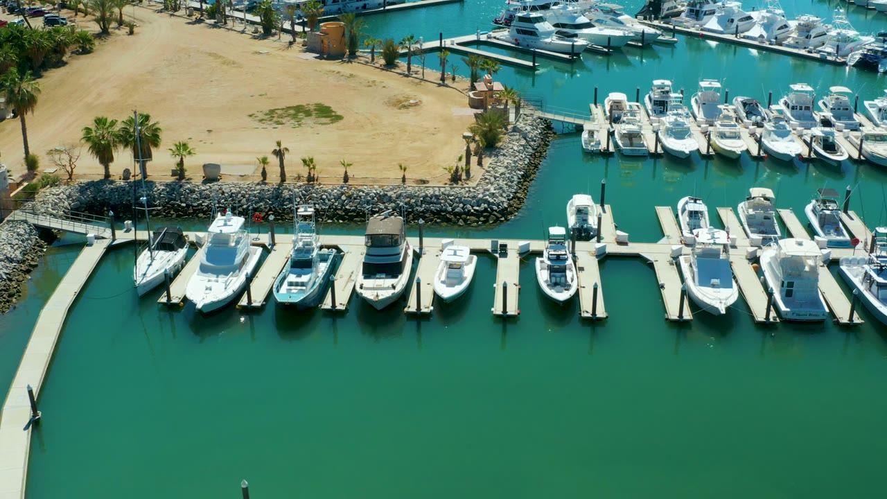 Overhead marina scene with docked boats, pale piers, turquoise water, and sandy land behind.