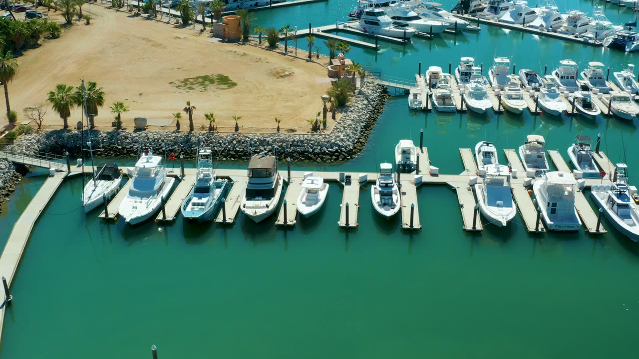 Puerto Los Cabos aerial frame with open green-blue marina water in the foreground, a line of docked boats across the center, sandy shore at the top, and no visible people.