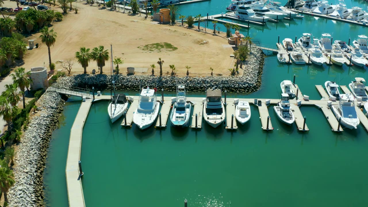 Aerial view of Puerto Los Cabos marina with white boats along docks, green water, a stone breakwater, sandy ground, palm trees, and a small person visible on the left dock.