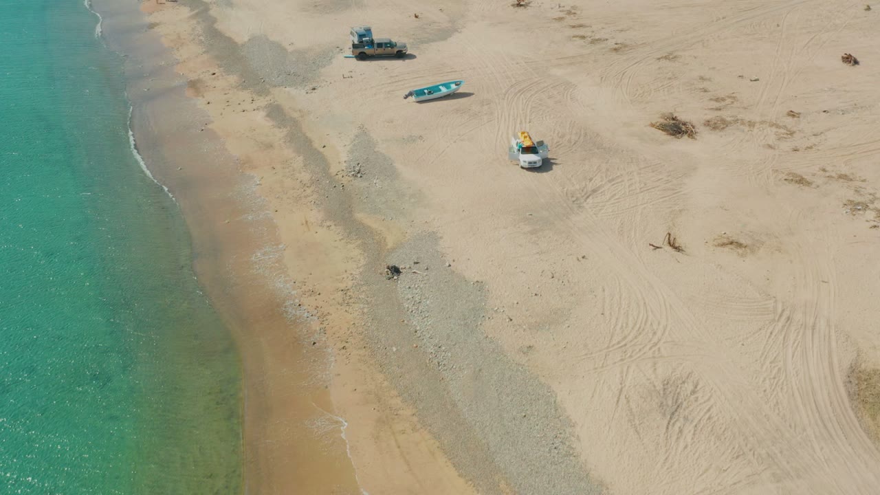 Aerial frame from Los Frailes showing more sand to the right, turquoise water at left, a parked truck above center, a small blue boat, a white vehicle or boat with yellow gear, tire marks, and no visible people.
