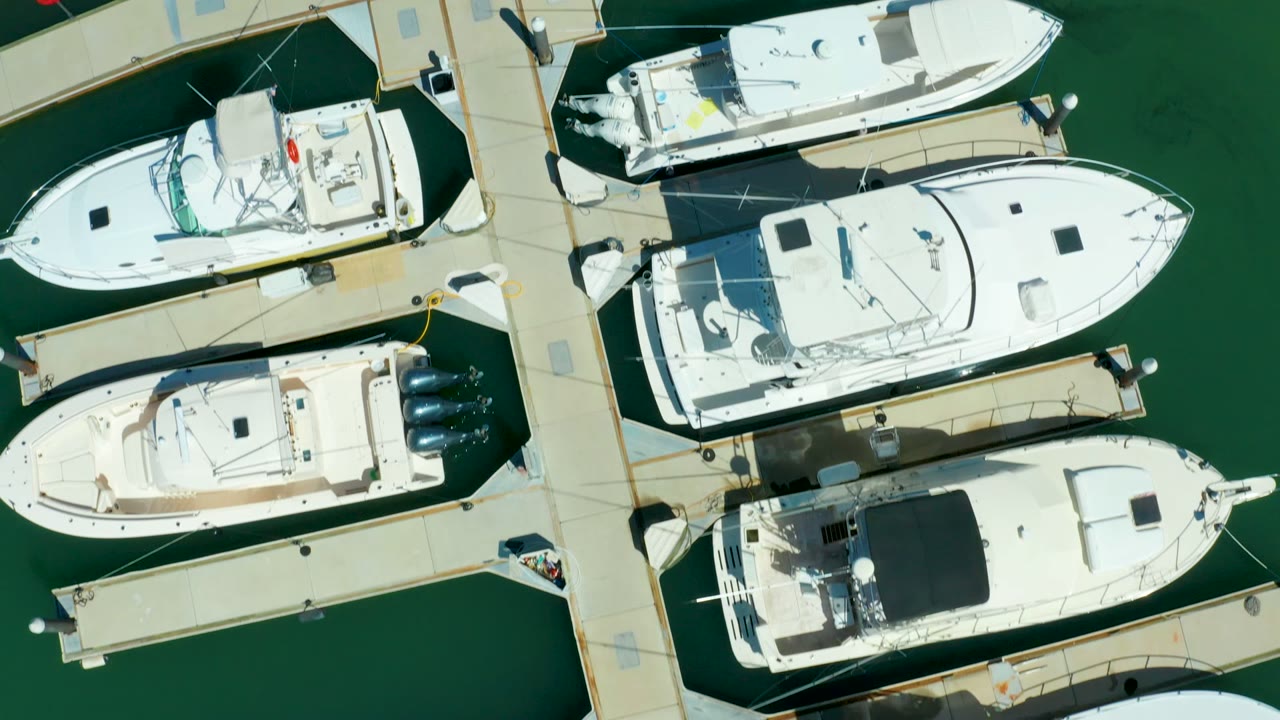 Overhead view of white boats docked along pale marina piers in Puerto Los Cabos, with green water and no visible people.