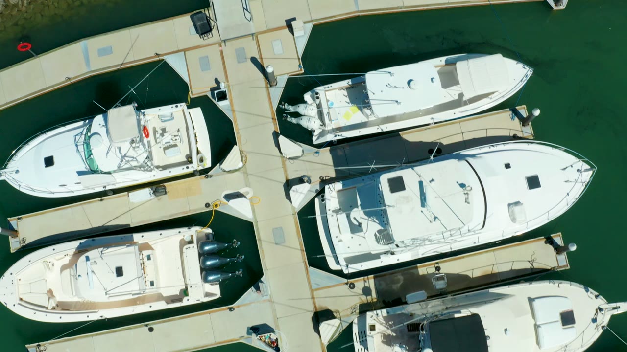 Top-down view of moored white boats at Puerto Los Cabos, with a central walkway, calm green water, and no visible people.