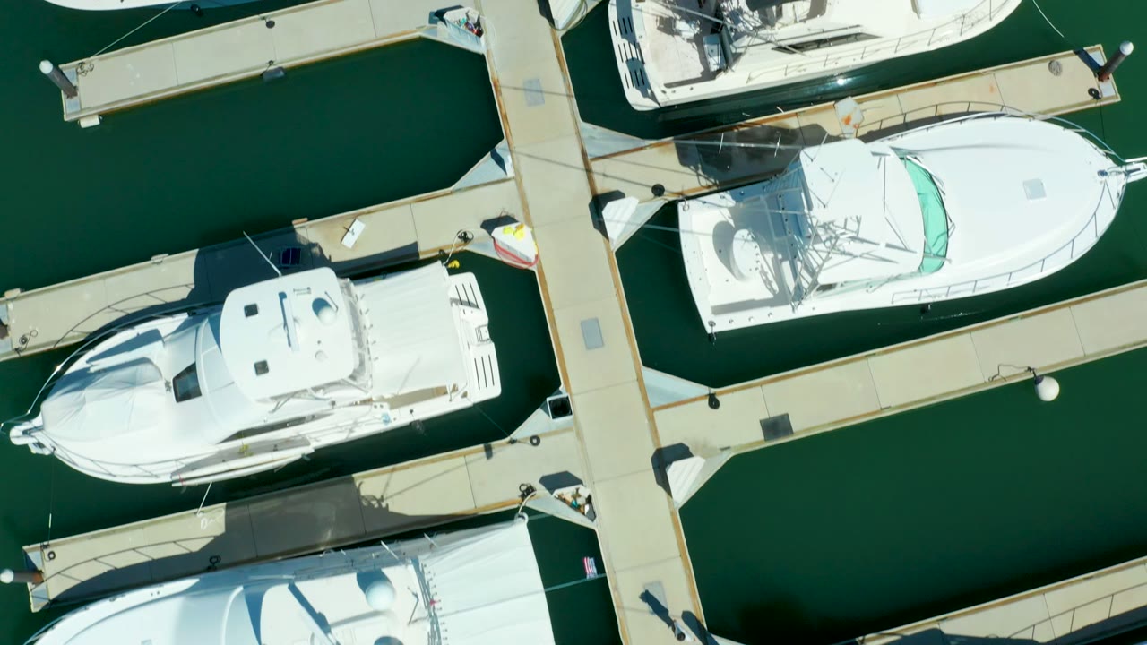 Top-down aerial frame over Puerto Los Cabos marina docks, with white boats beside pale dock fingers, green water channels, sharp shadows, and no visible people.