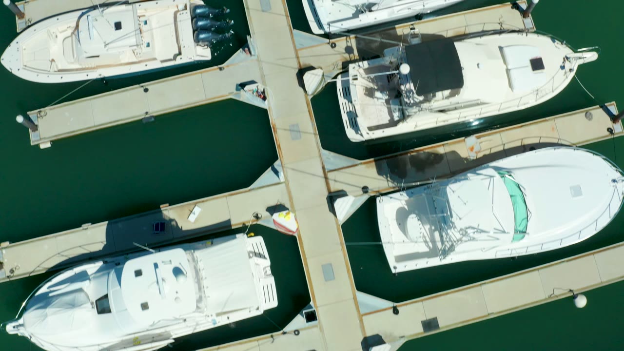 Overhead Puerto Los Cabos marina frame with white watercraft on both sides of pale docks, green water gaps, outboard motors, and no visible people.