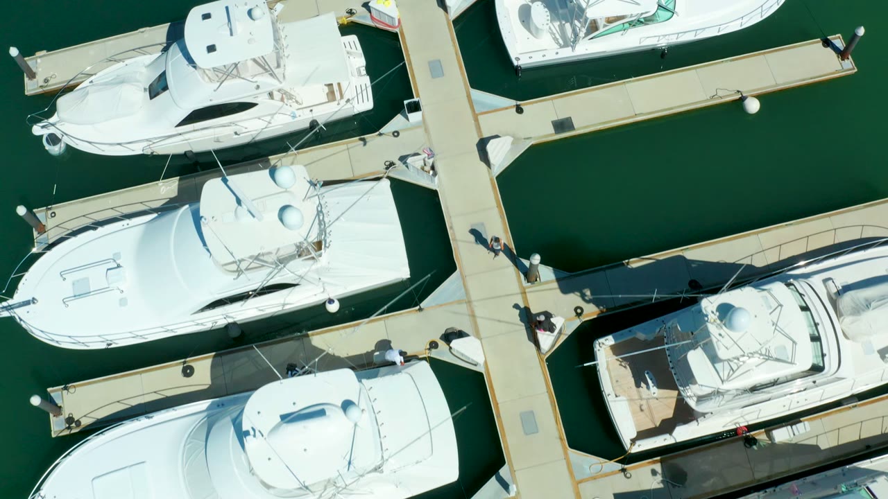 Overhead view of Puerto Los Cabos marina with several white yachts tied along tan piers, green water between slips, and small people visible on the docks.