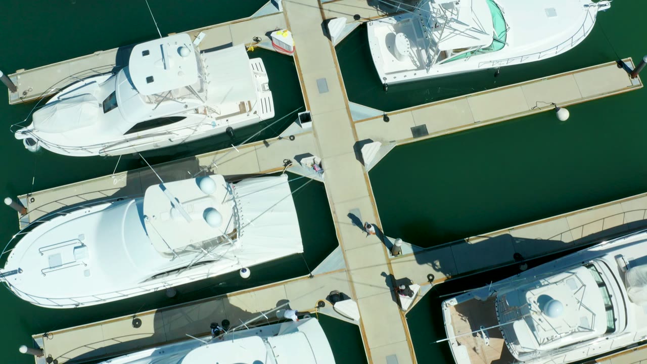 High overhead frame of Puerto Los Cabos marina with white boat roofs, finger piers, calm green water, and a few small people visible near the dock intersections.