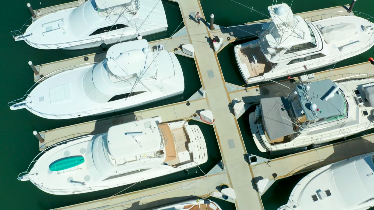 Overhead frame of white yachts moored around pale docks in Puerto Los Cabos, with green marina water and small people visible on the dock near the upper right.