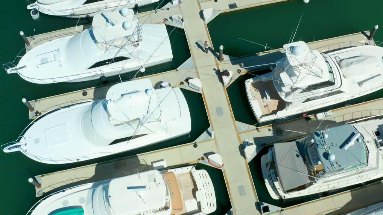 Puerto Los Cabos marina seen from above with bright white yacht roofs, a darker cockpit cover on one boat, narrow docks, green water, and people walking along the dock.