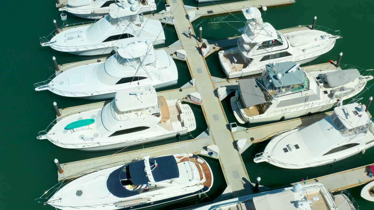 Overhead frame at Puerto Los Cabos showing white yachts moored on both sides of a central dock over green water, with small people visible on the pier near the upper right.