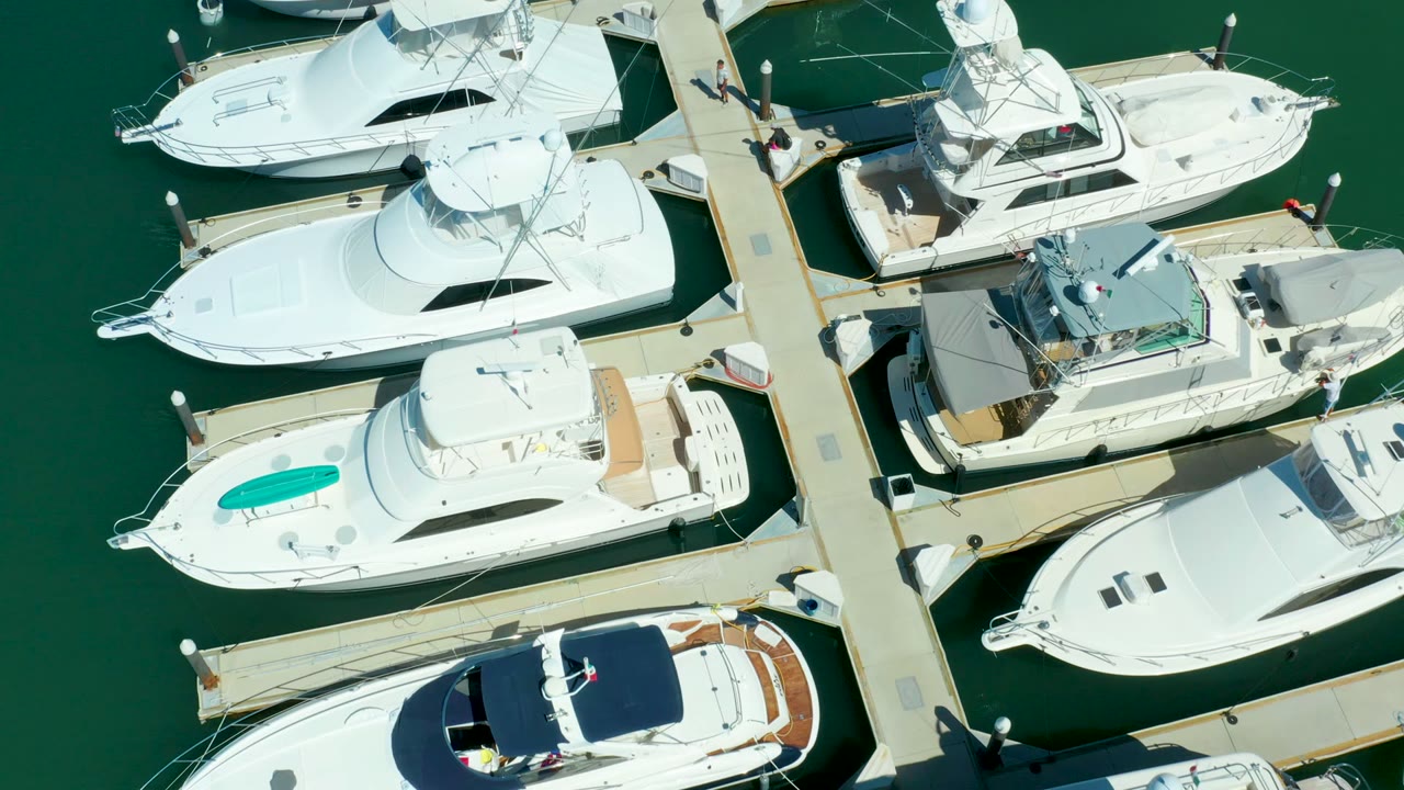 Overhead frame at Puerto Los Cabos showing a cluster of docked white yachts, a straight marina walkway, green water, and small figures on the dock near the right side.