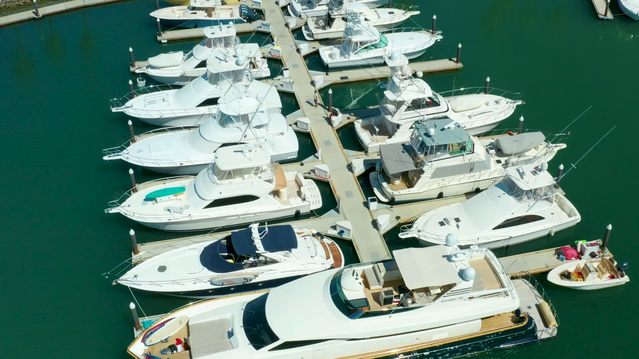 Overhead frame of Puerto Los Cabos marina with white yachts moored beside a central pier, deep green water, and no visible people.