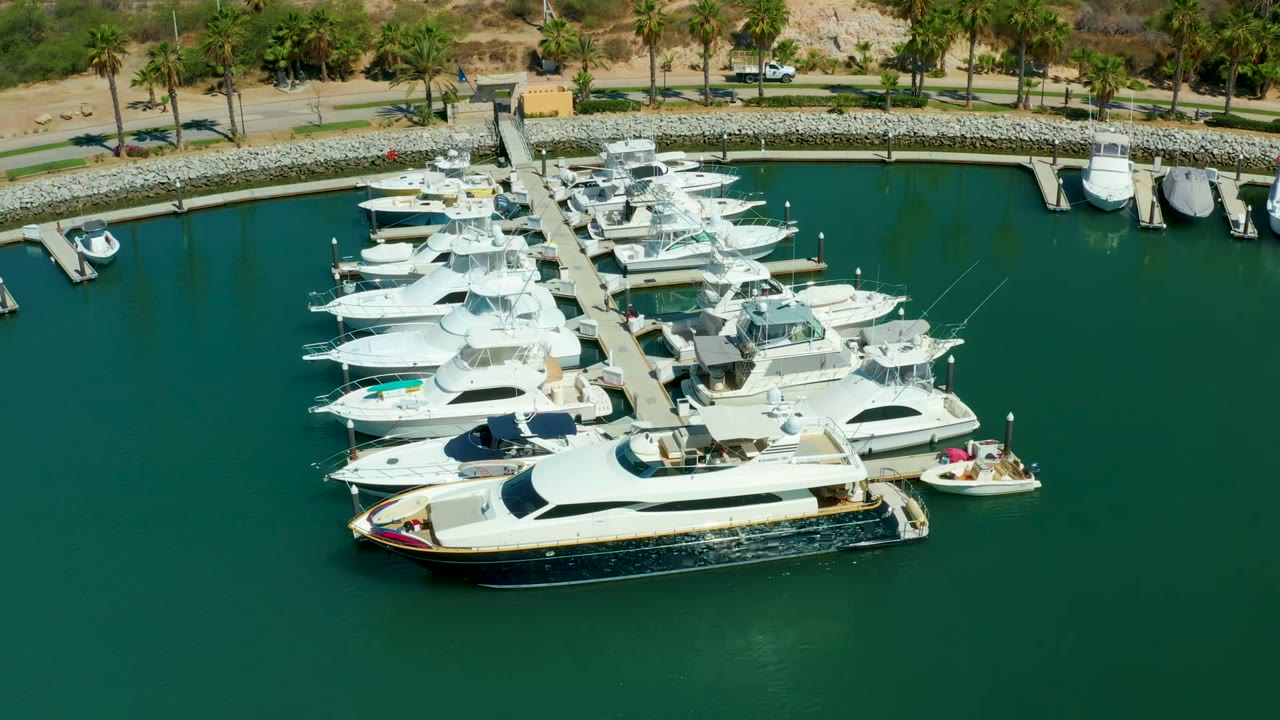 Aerial view over Puerto Los Cabos marina with white yachts along a central dock, green water, a shoreline road, palm trees, and no visible people.