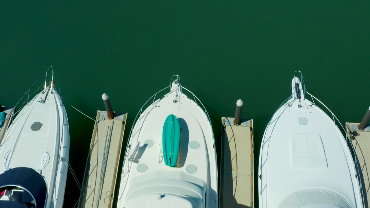 Overhead frame from Puerto Los Cabos showing white boats lined along dock fingers in dark green marina water, with teal deck covers visible and no visible people.
