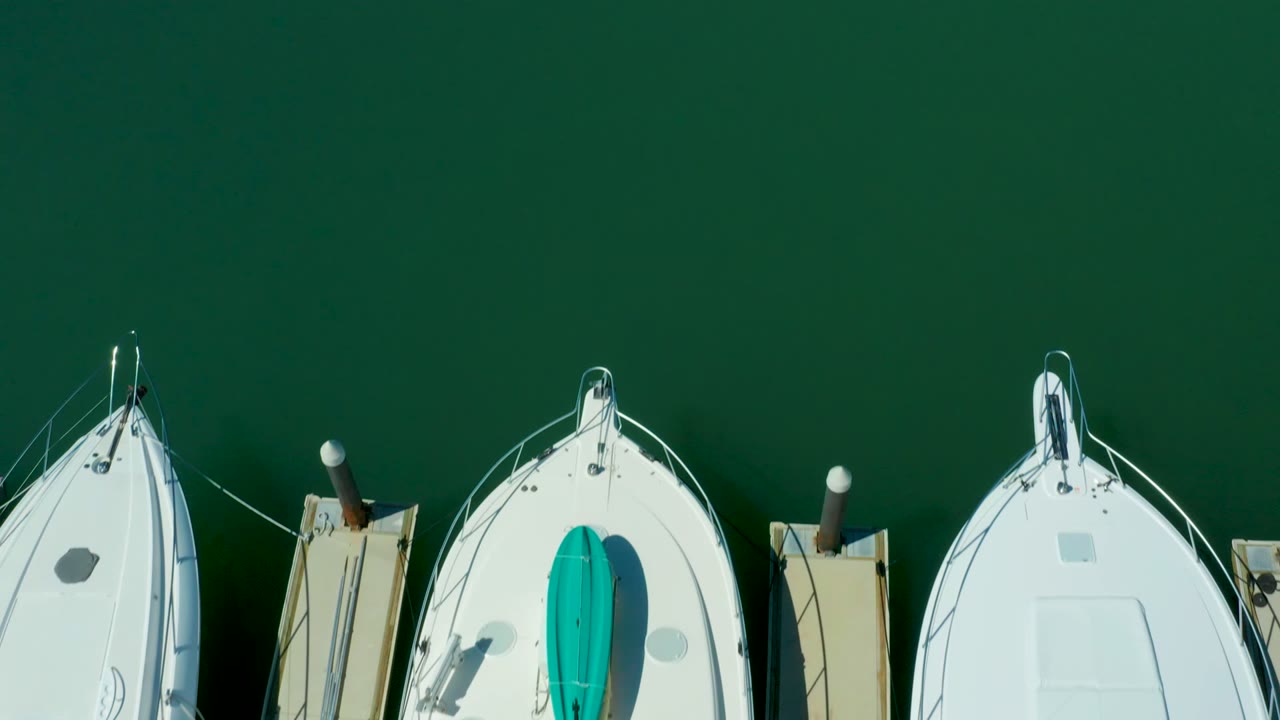 At two seconds, the aerial view in Puerto Los Cabos places white boat decks and dock posts along the lower edge, leaving a wide field of green marina water above them and no visible people.