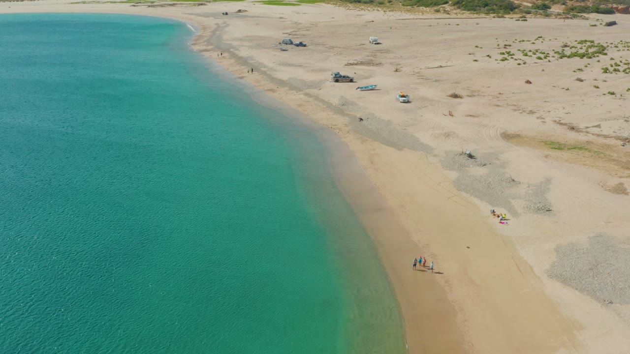 Aerial frame over Los Frailes, Los Cabos, showing turquoise water along the left edge, a sandy beach, small people near the lower shoreline, and vehicles set back on the sand.