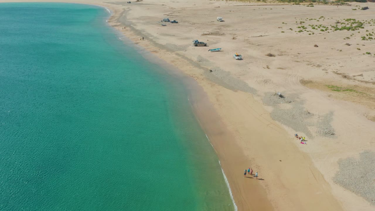 Drone view of clear water and sandy shore with beachgoers, vehicles, and a small boat inland.