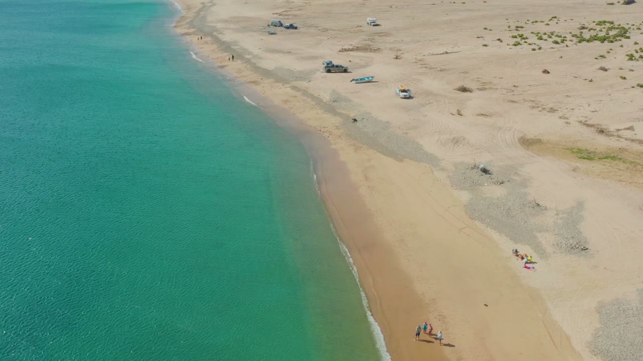 High aerial frame from Los Frailes showing a long strip of turquoise water, a narrow white wave line, open sand, small people near the lower right, and vehicles spread across the upper beach.