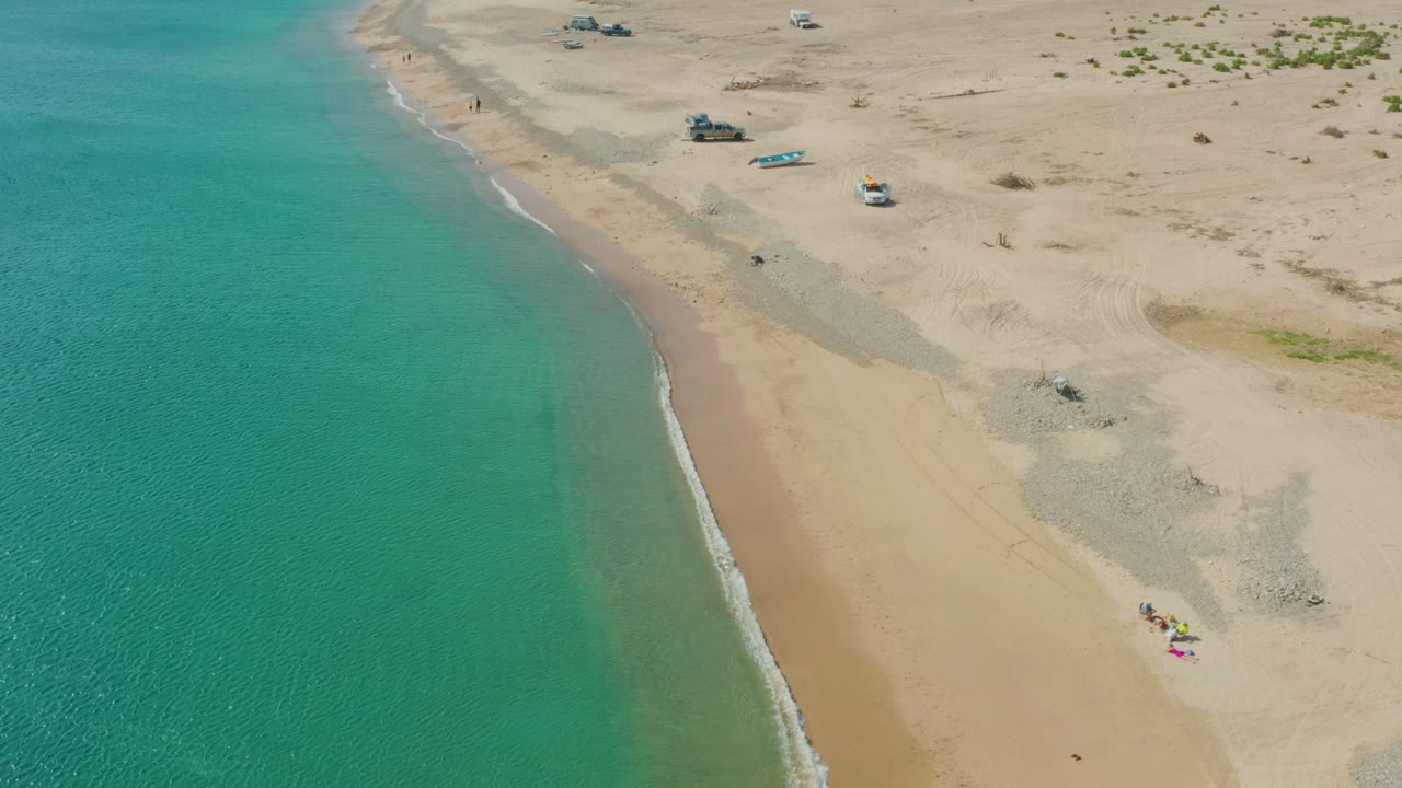Aerial shoreline with foamy surf, turquoise water, sandy beach, people, and parked vehicles.