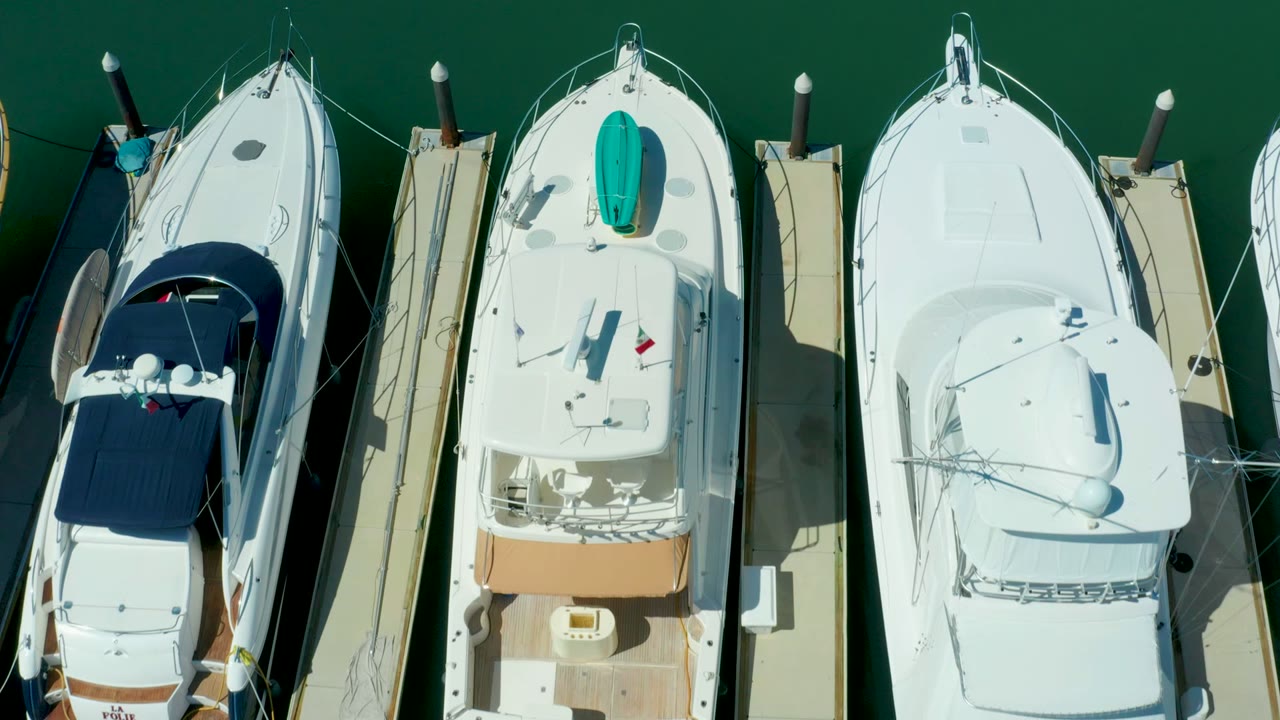 Overhead frame from Puerto Los Cabos showing white boats moored beside narrow wooden docks in green marina water, with dark canvas covers and no visible people.