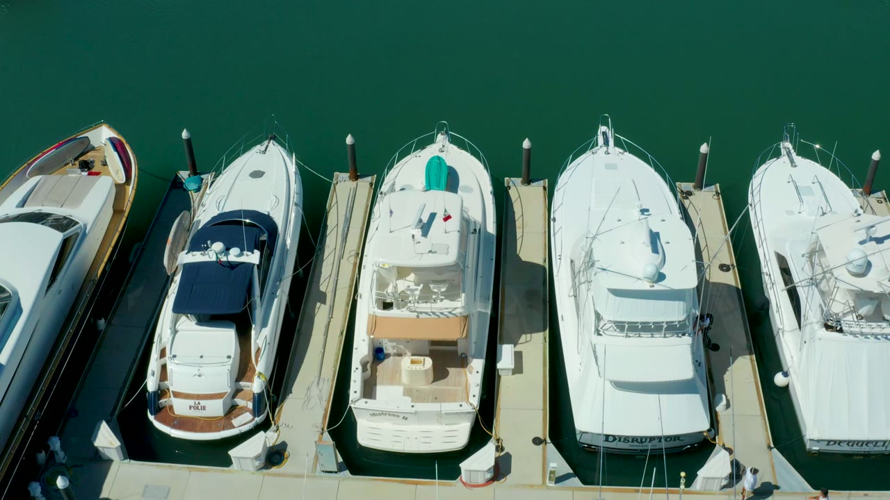 Opening overhead frame of docked white boats in Puerto Los Cabos, Baja California Sur, with pale slips, green water, blue canvas covers, and no visible people.