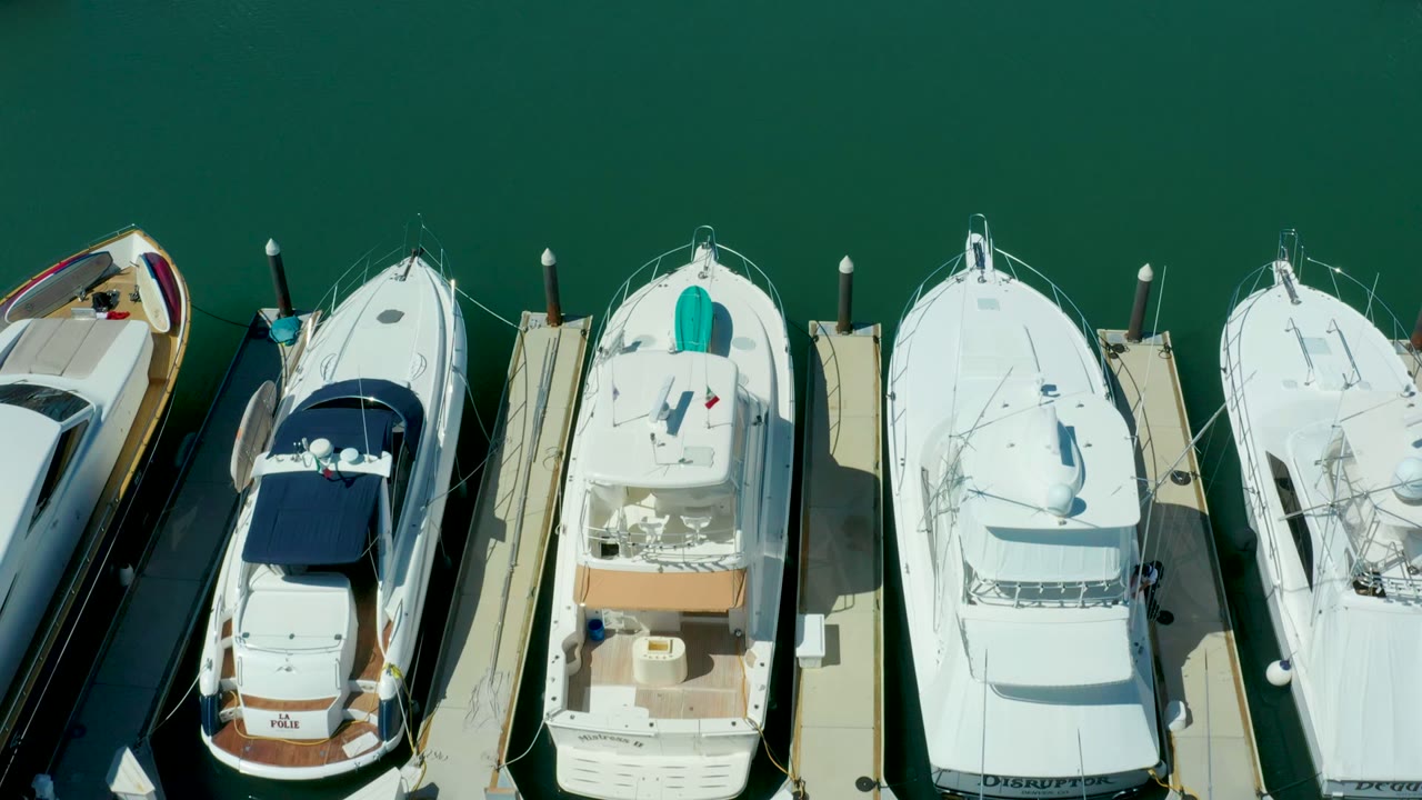 Overhead Puerto Los Cabos frame with docked boats separated by narrow walkways, visible fenders, bright white decks, green water, and no visible people.