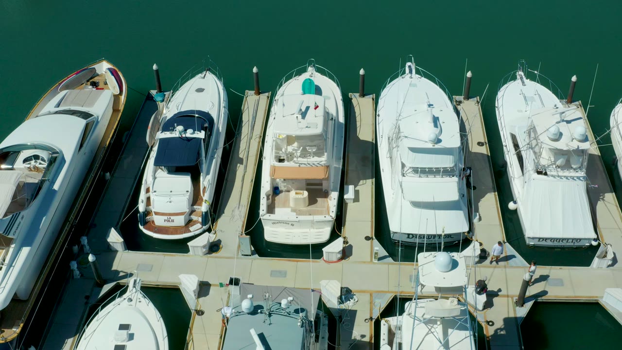 Overhead frame in Puerto Los Cabos showing white yachts and smaller boats moored along a pale marina dock, with green water above the slips and small people visible on the walkway.