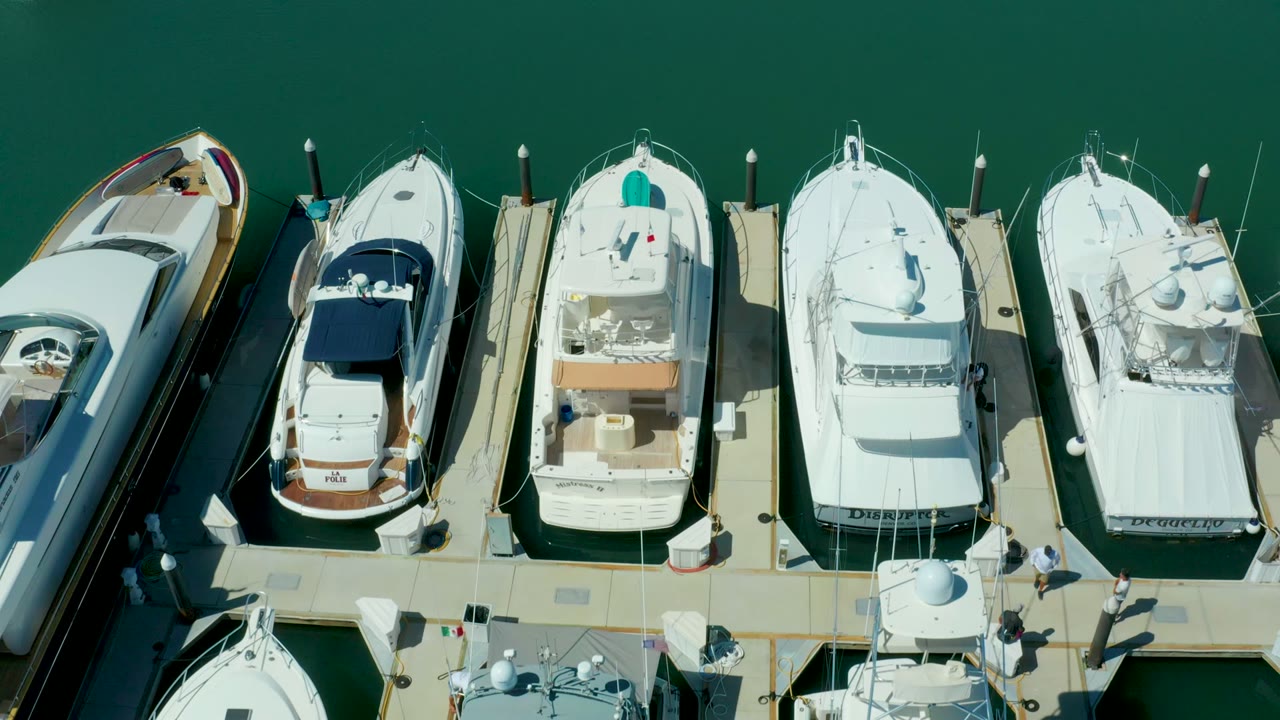 At 0:02, the overhead Puerto Los Cabos view shows docked white boats on both sides of the walkway, hard sunlight on the dock, and small people visible near the lower right pier area.