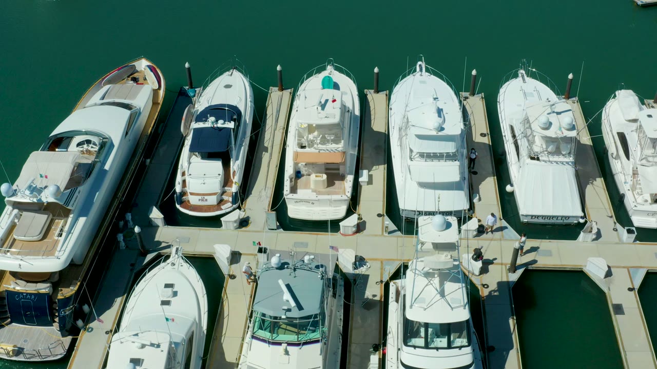 High overhead Puerto Los Cabos marina frame with several white yachts, a tan floating pier, dark dock shadows, calm green water, and no visible people.