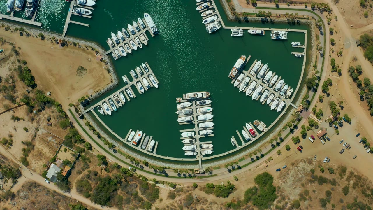 Overhead view of Puerto Los Cabos marina with white boats in green water, dry land beside a curved road, parked cars, sparse vegetation, and no visible people.