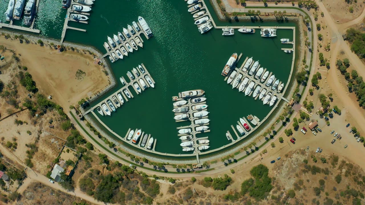 Top-down frame of Puerto Los Cabos marina showing several dock arms lined with boats, a curved basin edge, dry shoreline, small vehicles, sparse trees, and no visible people.