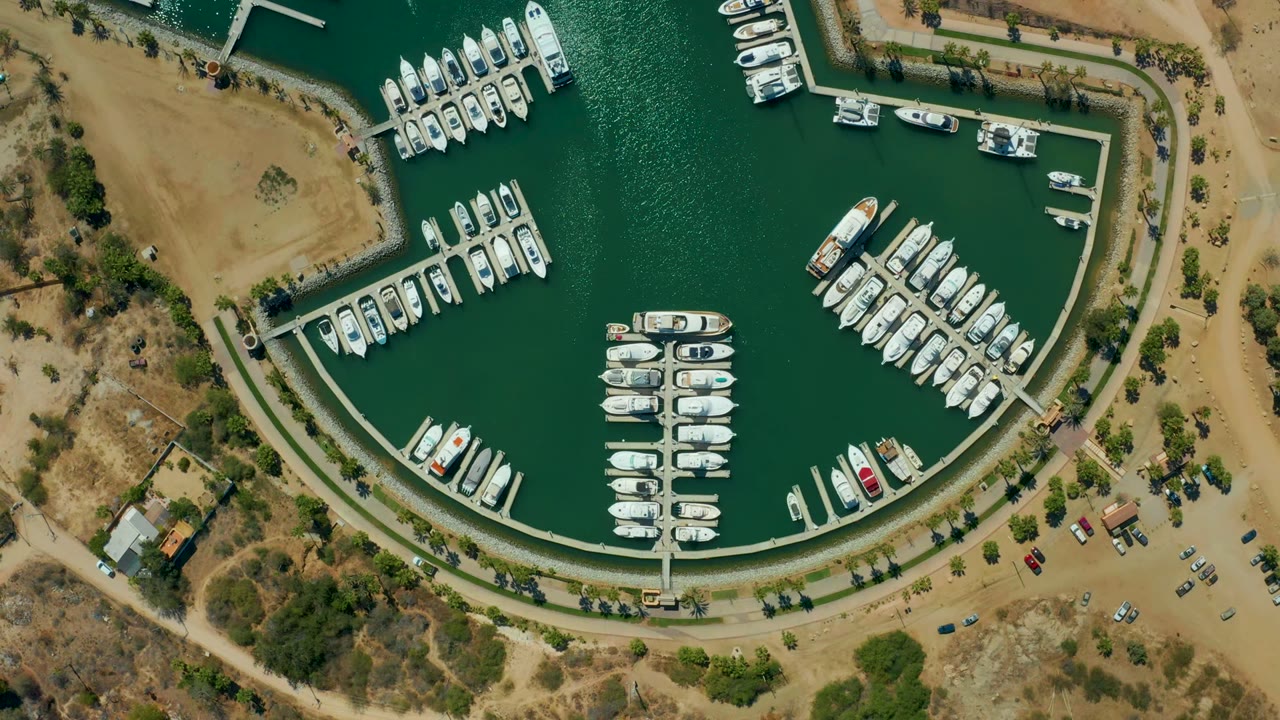 Top-down aerial frame of Puerto Los Cabos marina with white boats moored along curved docks, green water, dry shoreline, small roads, parked cars, and no visible people.