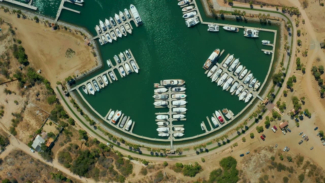 Third frame from the Puerto Los Cabos aerial shows moored white boats arranged around the marina basin, green water texture, tan undeveloped land, a road, parking area, and no visible people.