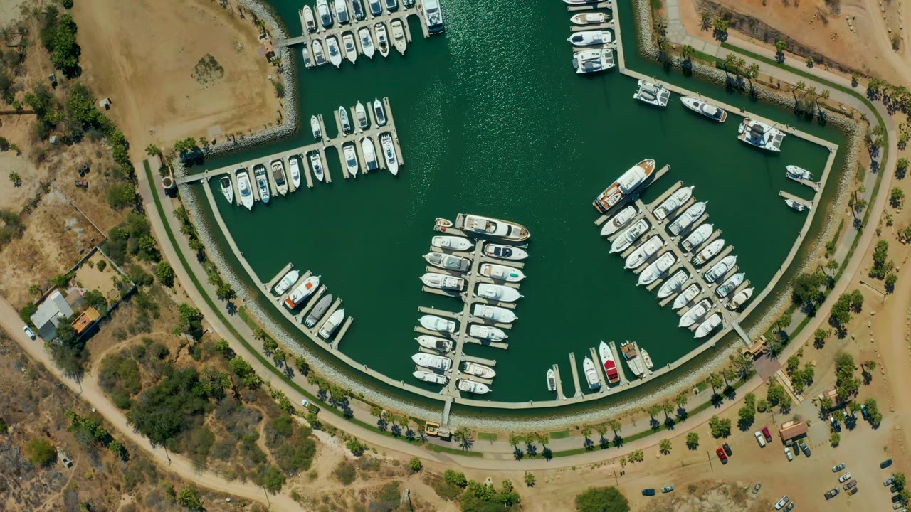 Overhead view of Puerto Los Cabos marina with rows of white boats, green water, concrete docks, dry shoreline, sparse trees, parked cars, and no visible people at the start of the clip.