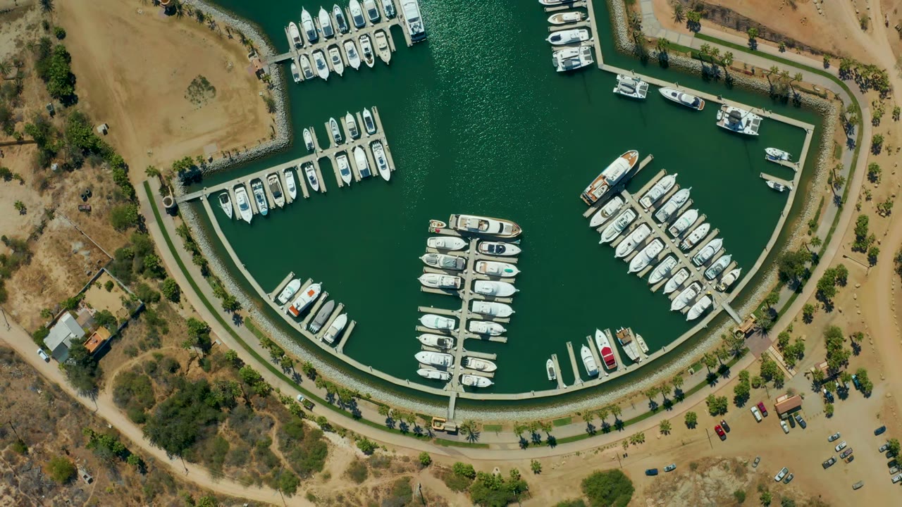 Mid-clip overhead frame in Puerto Los Cabos with rows of boats in rippled green water, docks crossing the basin, dry scrubland at the edges, parked cars, and no visible people.