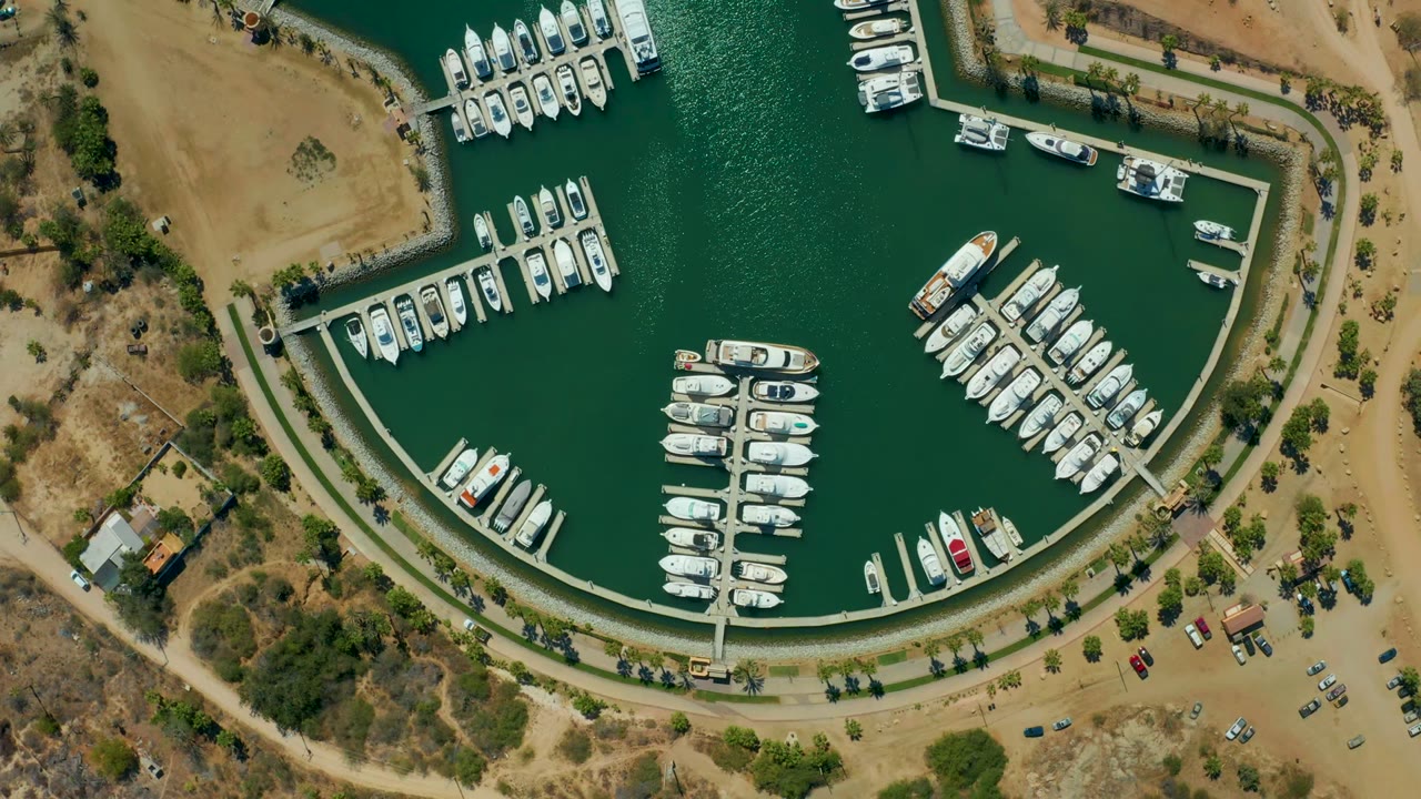 Drone view of a marina with boats, docks, dry shore, and parked vehicles.
