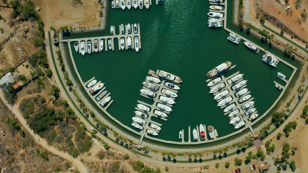 Overhead view of Puerto Los Cabos marina with green water, white docked boats, curved docks, dry land, a shoreline road, and no visible people at the start of the clip.