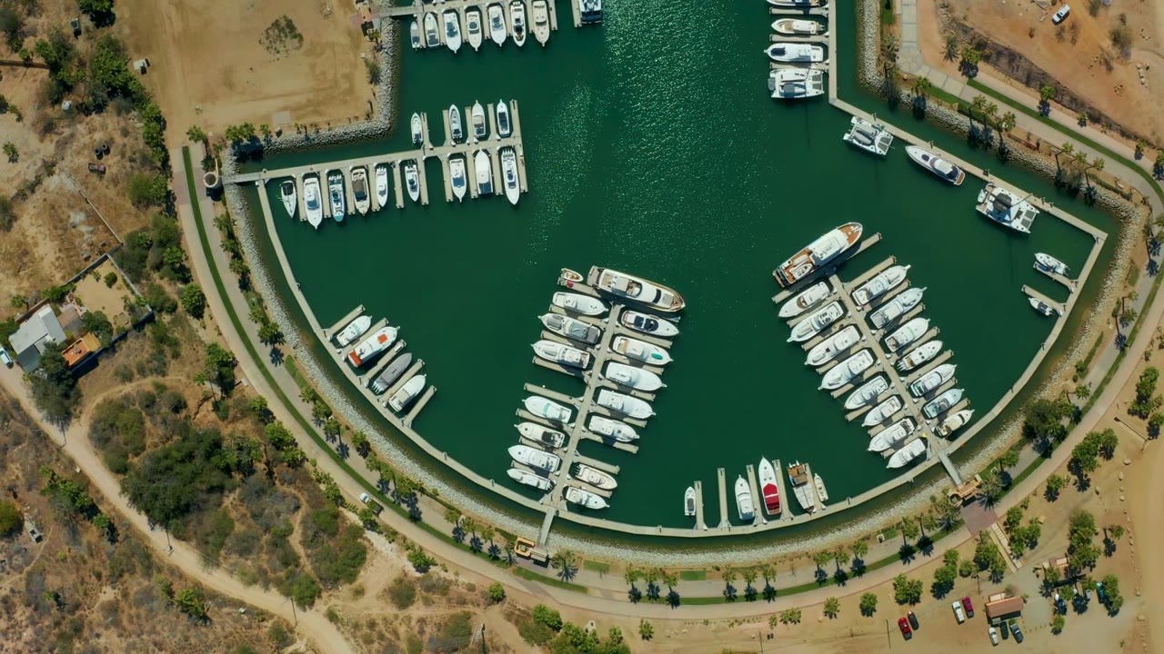 Aerial overhead frame of Puerto Los Cabos marina with two main clusters of docked boats, textured green water, dry lots, shoreline paths, cars near the road, and no visible people.