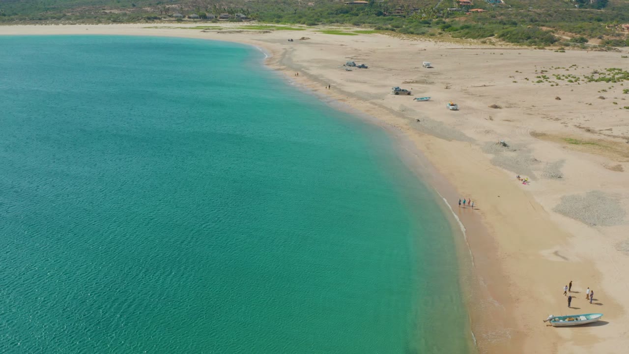 Aerial frame from Los Frailes, Baja California Sur, showing turquoise ocean along a curved sandy beach with small groups of people near the waterline and a boat on the lower right sand.