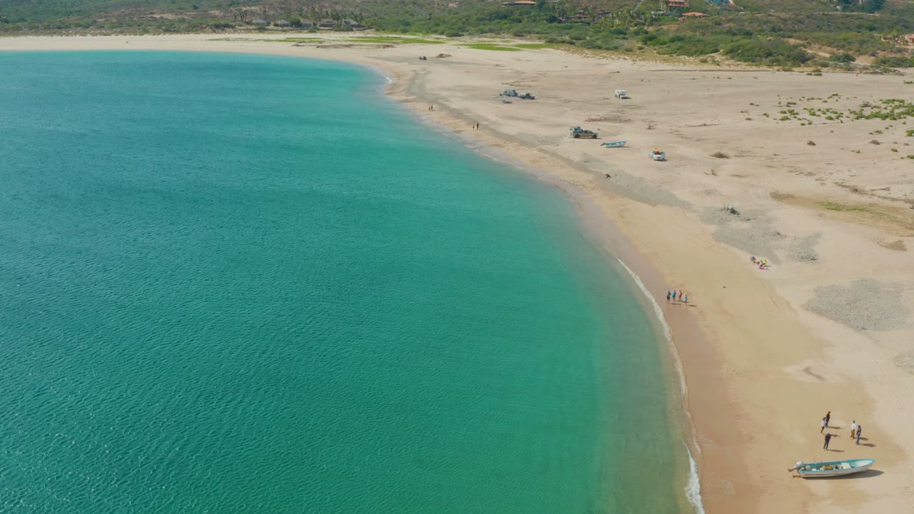 Wide aerial shoreline with clear water, sand, people, and boats on the beach.
