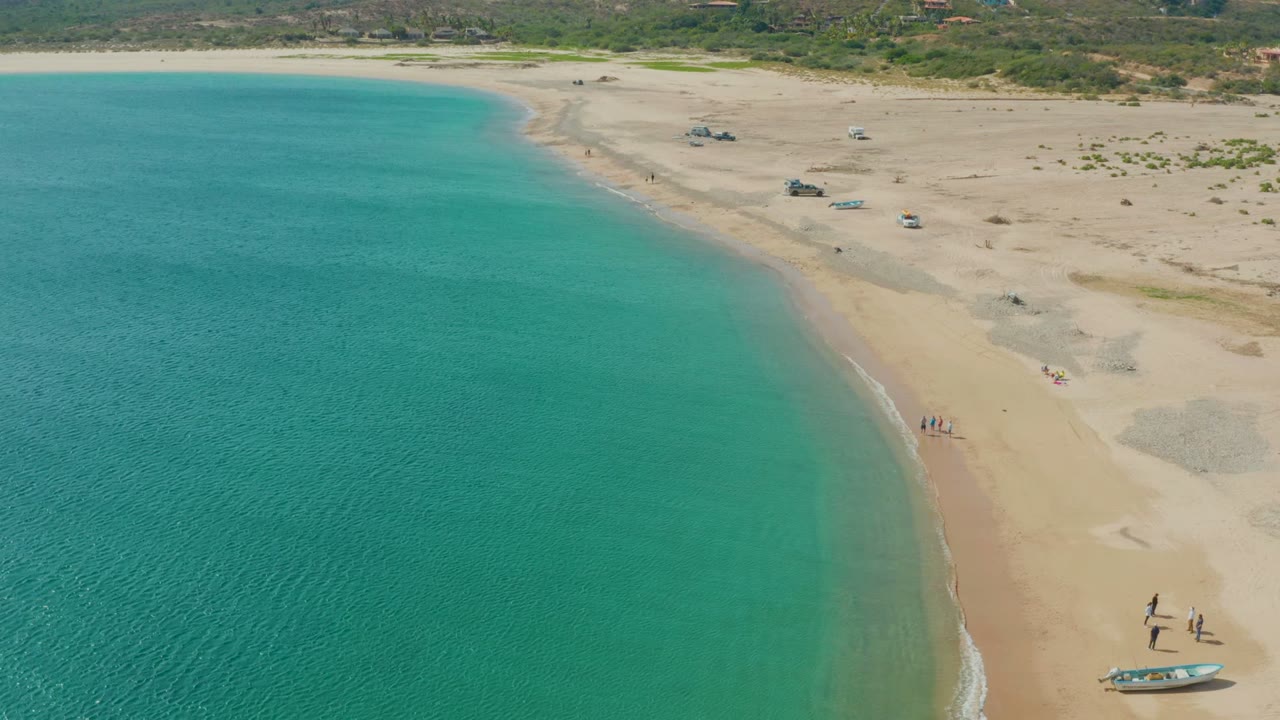 The Los Frailes shoreline curves through the frame, with clear turquoise water on the left, pale sand on the right, beachgoers near the wet sand, and small boats visible along the upper beach.