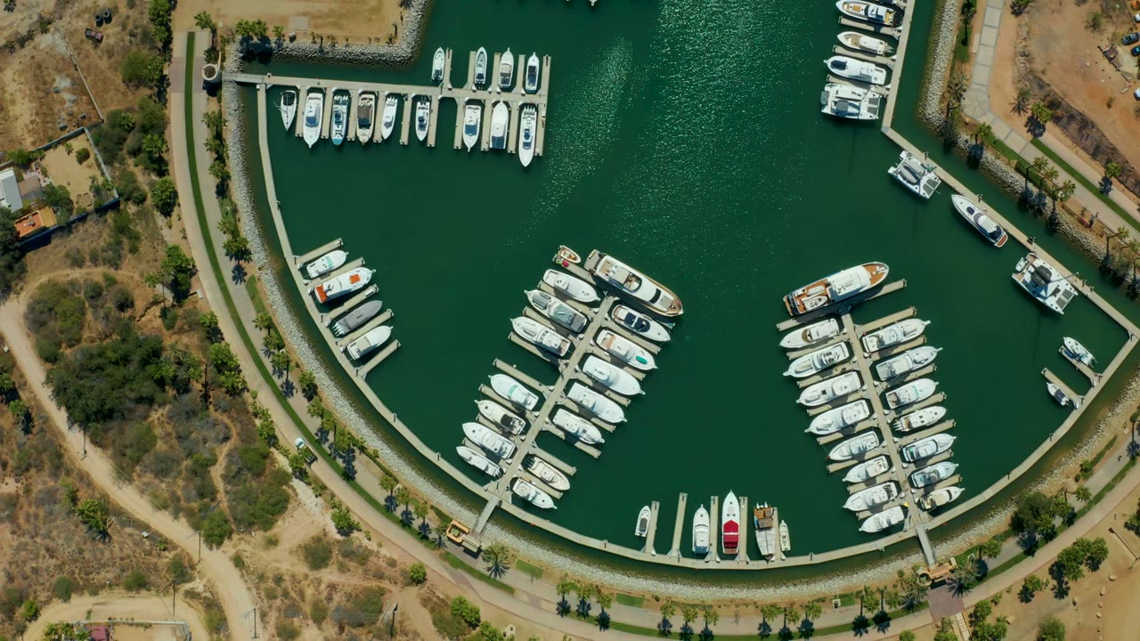 Overhead view of Puerto Los Cabos marina with rows of white boats, green water, a curved seawall path, dry land, sparse bushes, and no visible people.