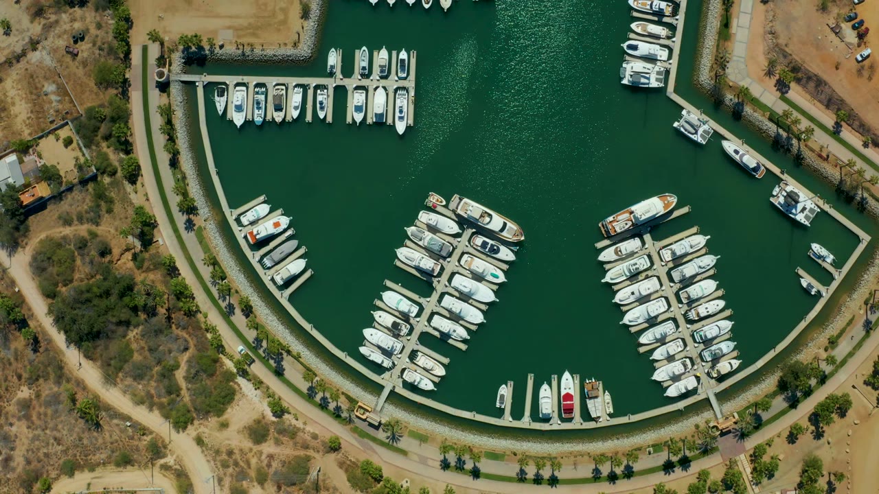 Puerto Los Cabos overhead marina view with multiple white boats at slips, a curved concrete edge, textured green water, dry shoreline, and no visible people.