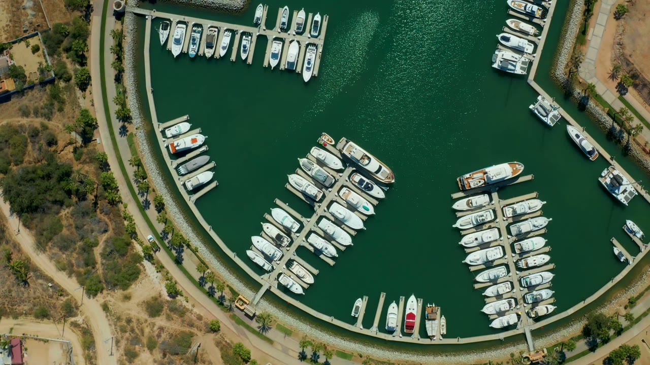 Overhead view of Puerto Los Cabos marina with white boats along curved docks, green water, tan shoreline paths, sparse vegetation, and no visible people.