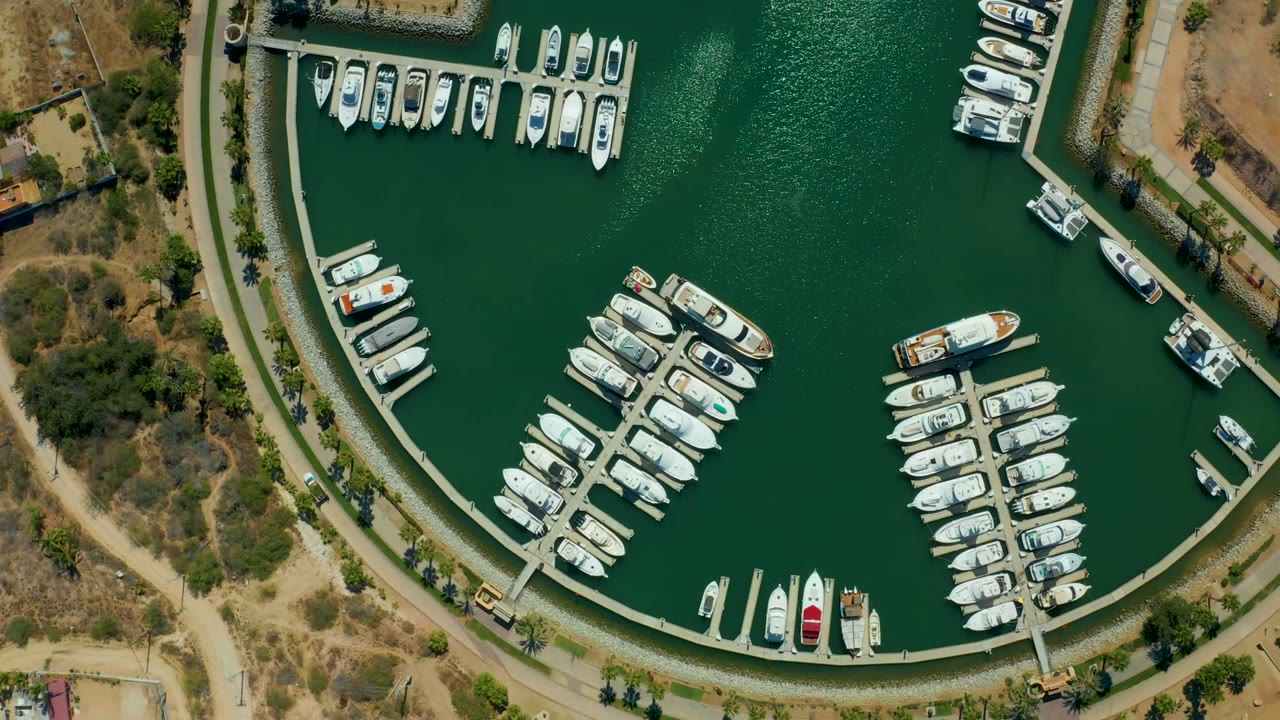 Drone view of Puerto Los Cabos marina with clustered yachts in slips, a broad green water channel, dry shoreline terrain, small structures, and no visible people.