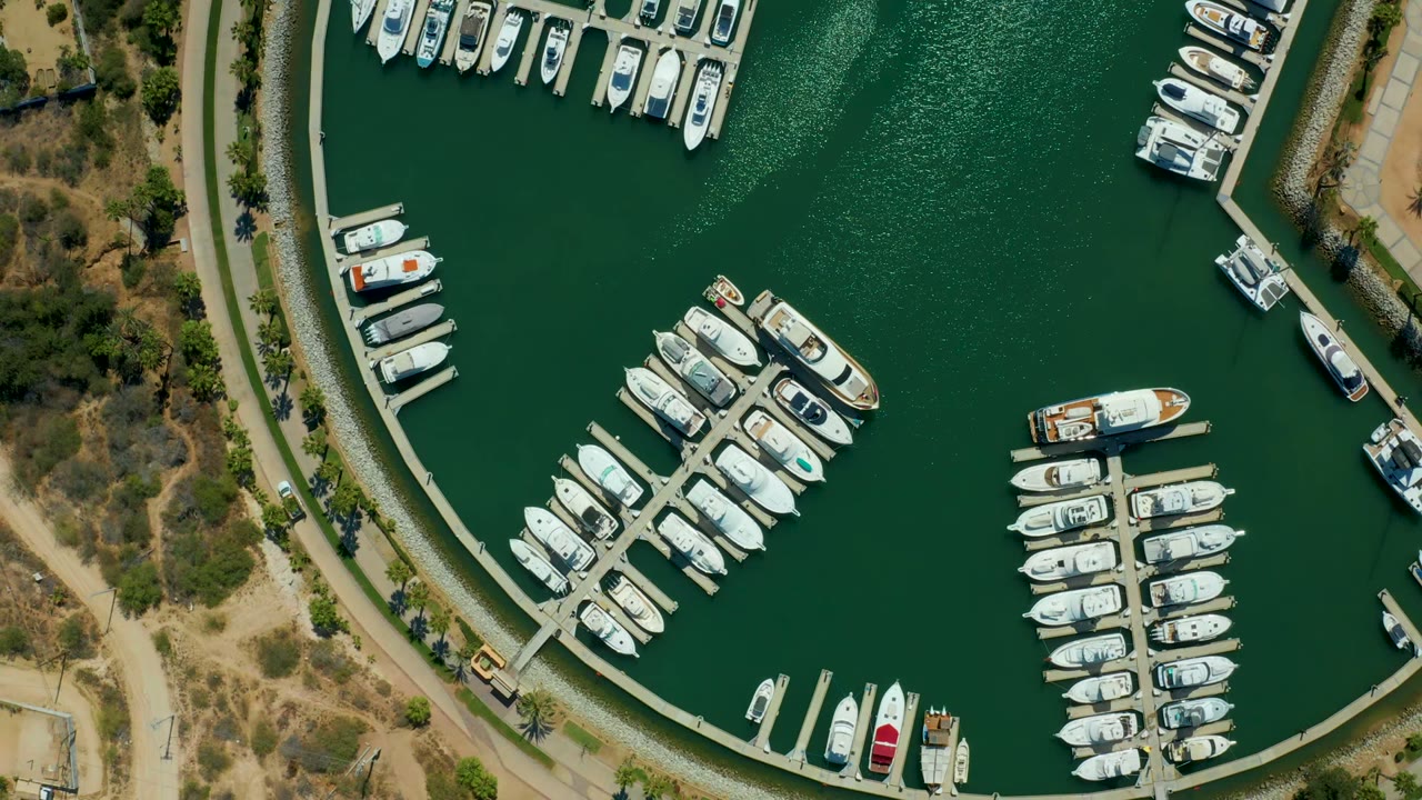 Opening overhead frame of Puerto Los Cabos marina showing curved docks, clustered white boats, green water, dry shoreline paths, and no visible people.